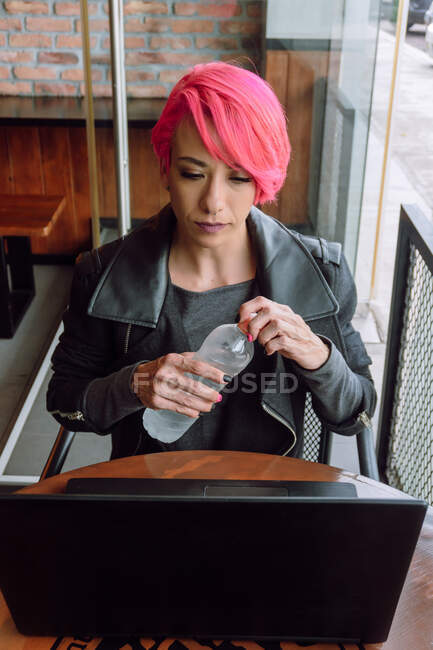 Elegante giovane donna con i capelli tinti in abito nero seduto a tavola e avere una bottiglia d'acqua mentre si guarda lo schermo del computer portatile in caffè — Foto stock