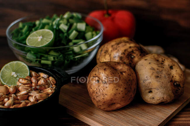 Alto ângulo de batatas cruas e cebolinha picada colocados em tábua de corte de madeira perto da tigela com grãos preparados para cozinhar caldo de galinha — Fotografia de Stock