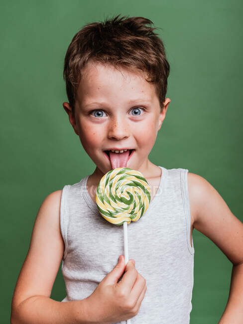 Preteen boy licking tasty spiral lollipop on green background in studio while looking at camera — Stock Photo