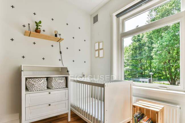 White crib placed between cabinet and bookshelf near window in sunlit nursery at home — Stock Photo