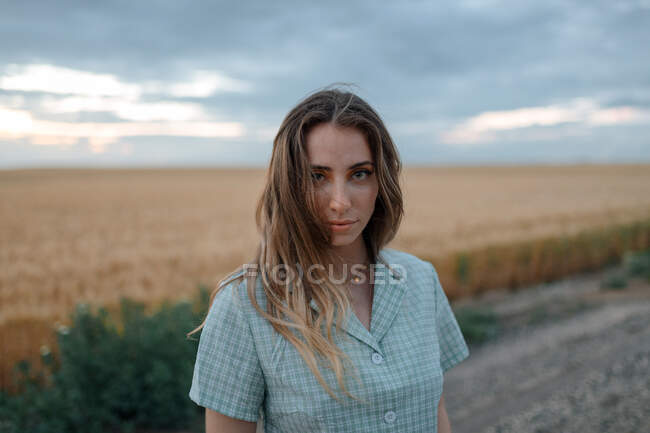 Jeune femme consciente regardant la caméra sur la route près de prairie sous le ciel nuageux en soirée dans la campagne — Photo de stock