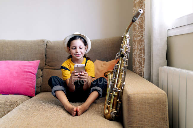 Kind mit Hut SMS auf Handy, während es mit Saxophon auf Couch im Wohnzimmer sitzt — Stockfoto