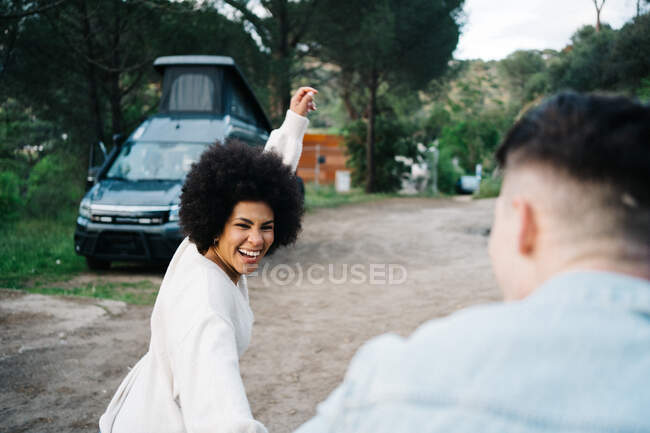 Femme afro-américaine souriante tenant la main du petit ami cultivé tenant la main tout en se promenant et en regardant loin contre le camping-car — Photo de stock