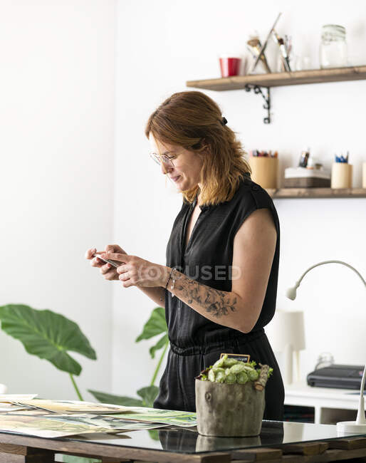 Веселая женщина-дизайнер фотографирует картины на смартфоне, стоя рядом со столом и работая в творческом рабочем пространстве — стоковое фото