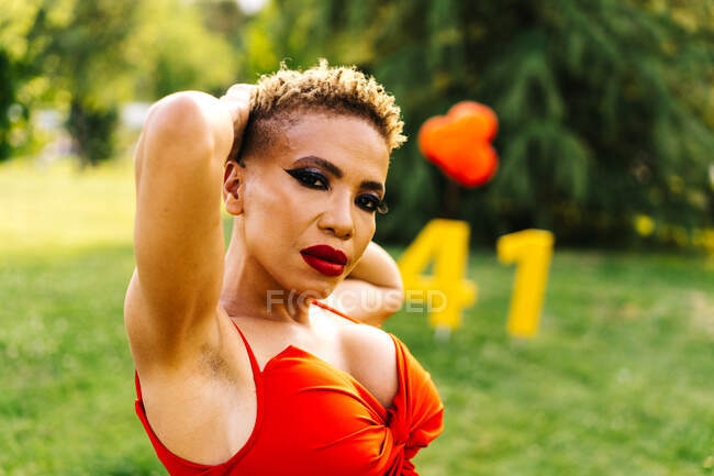 Trendige ethnische Frau mittleren Alters in roter Kleidung mit modernem Haarschnitt und Händen hinter dem Kopf, die während einer Geburtstagsfeier in die Kamera schauen — Stockfoto