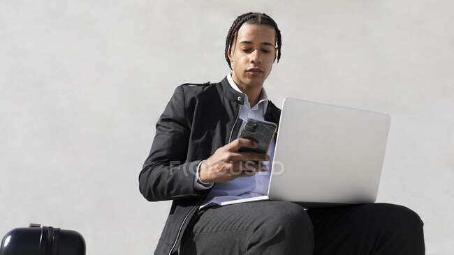 Молодой хорошо одетый этнический предприниматель мужчина с косичками просматривает на мобильном телефоне во время работы на ноутбуке сидя у стены на улице — стоковое фото