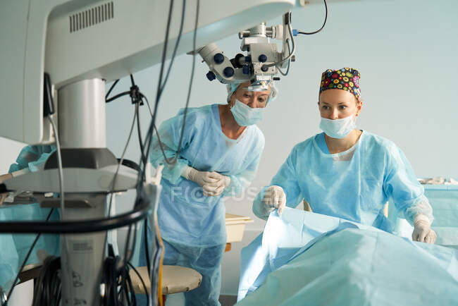 Doctora atenta en uniforme estéril contra su compañero de trabajo que mira hacia otro lado mientras se prepara para la cirugía en el hospital con microscopio - foto de stock