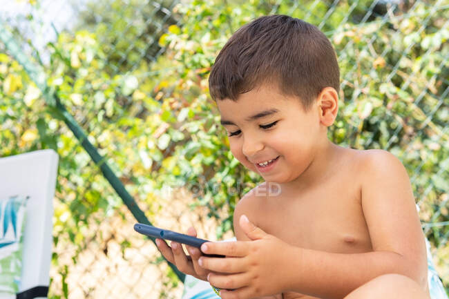 Alegre lindo niño usando el teléfono inteligente mientras está sentado en el jardín cerca de la cerca verde en el día de verano soleado - foto de stock