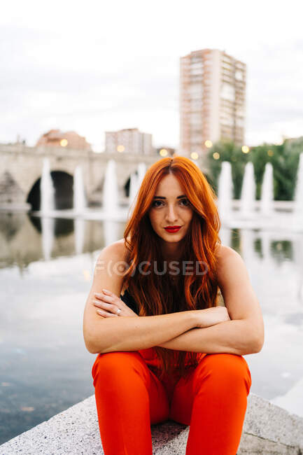 Красивая женщина с длинными рыжими волосами и в ярко-оранжевых брюках сидит на границе на набережной города и смотрит в камеру — стоковое фото