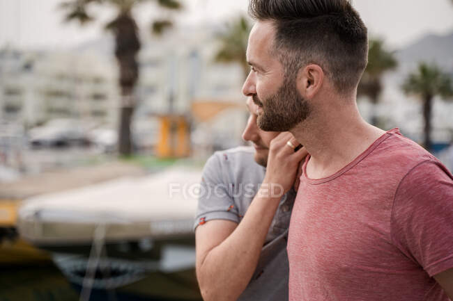 Conteúdo casal de homens homossexuais em t-shirts abraçando enquanto olha para longe no porto — Fotografia de Stock
