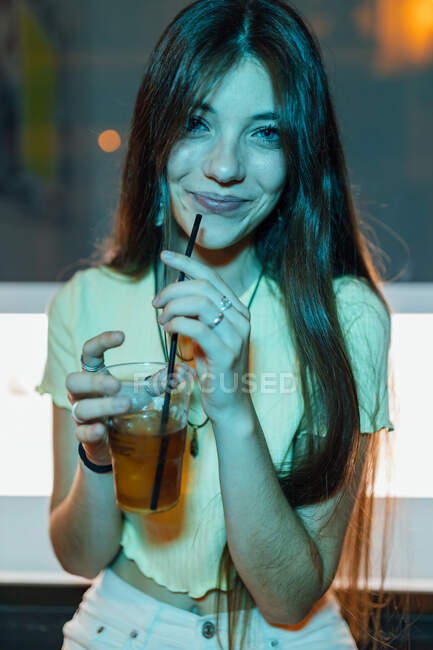 Contenu jeune femme aux cheveux longs et verre de boisson rafraîchissante en regardant la caméra au crépuscule — Photo de stock