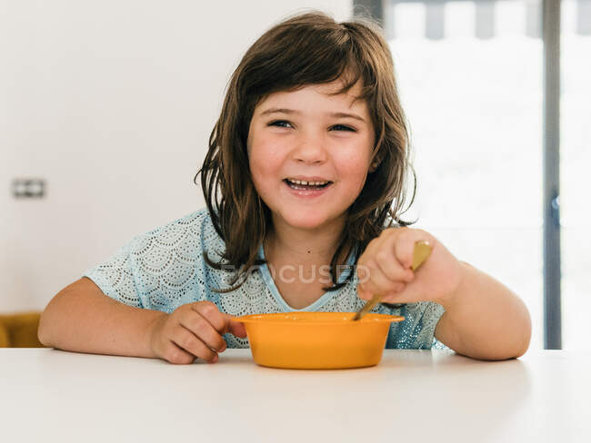 Очаровательный ребенок ест вкусный кремовый суп из пластиковой миски, сидя за столом во время обеда дома — стоковое фото