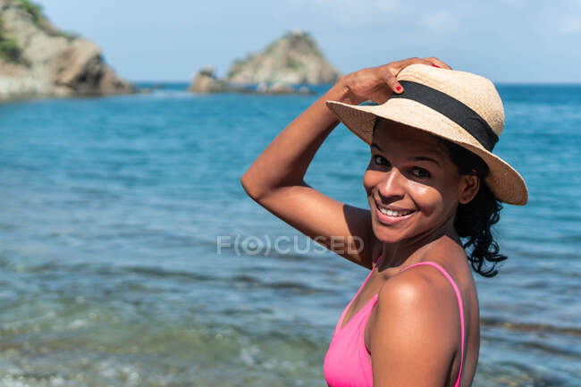 Vista lateral del alegre turista étnico femenino en traje de baño y sombrero mirando a la cámara en la costa del océano - foto de stock