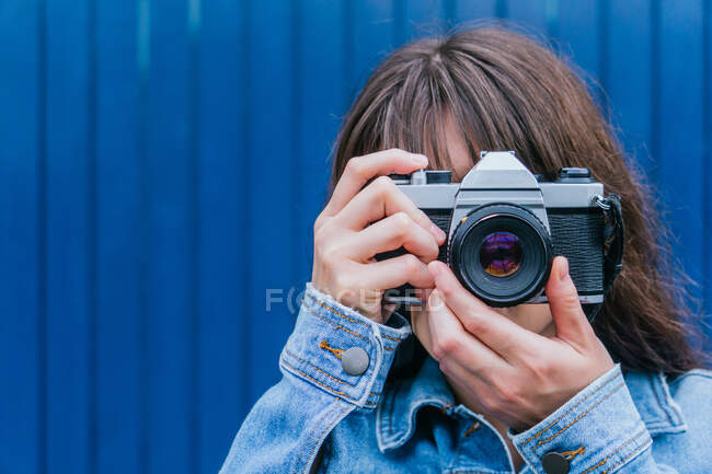 Fotógrafo feminino anônimo em jaqueta de ganga tirando foto na câmera de fotos vintage no fundo da parede azul na rua da cidade — Fotografia de Stock
