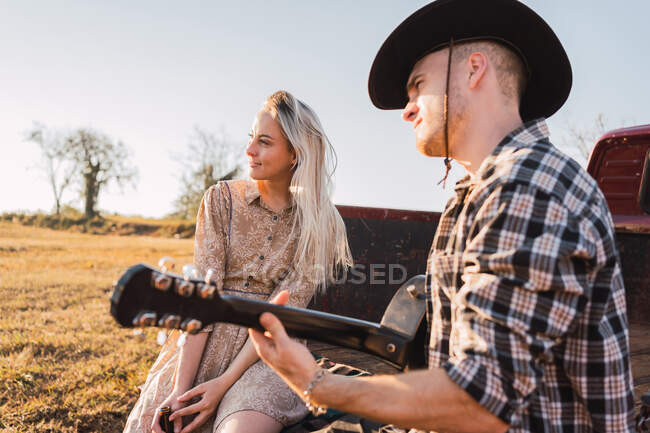 Freund mit Cowboyhut spielt Akustikgitarre, während er mit Freundin im Kofferraum eines roten Retro-Pickups sitzt, der auf einer sandigen Straße im Grünen geparkt ist — Stockfoto