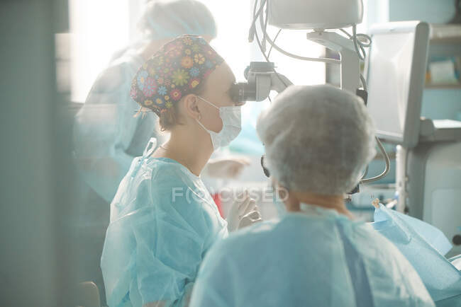Доросла жінка-лікар у стерильній масці та декоративній медичній кепці, яка дивиться через хірургічний мікроскоп проти колеги по сільськогосподарській галузі в лікарні — стокове фото