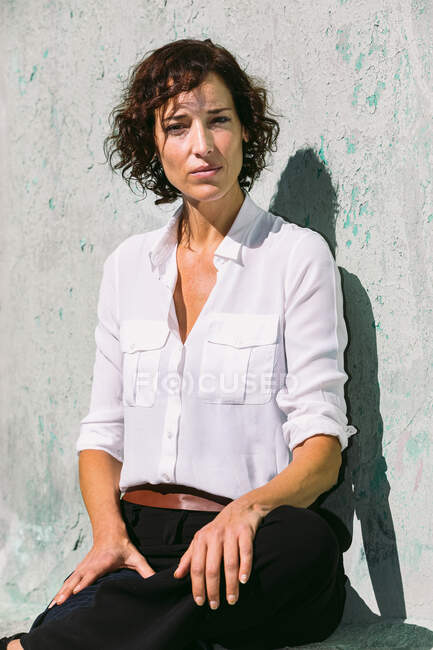 Задумчивая женщина в элегантной одежде стоит солнечный свет на потрепанной стене и смотрит в камеру — стоковое фото