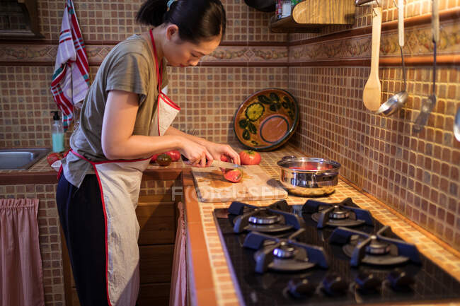 Vista lateral de la hembra étnica en delantal cortando tomates maduros en la tabla de cortar mientras se cocina el almuerzo en la cocina en casa - foto de stock