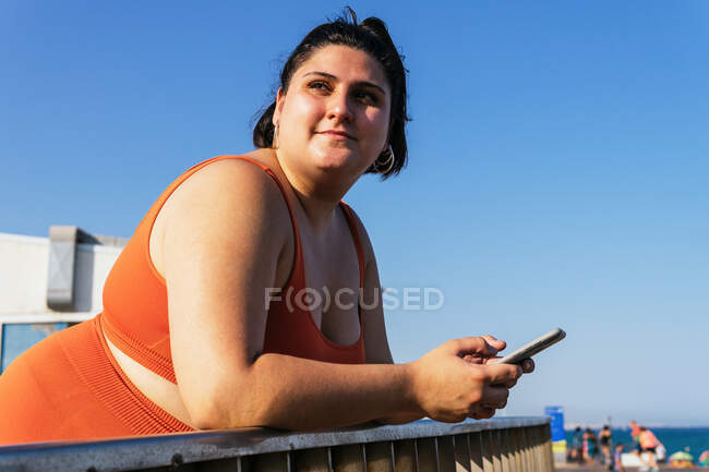 Мечтательная этническая спортсменка с пышным телом и мобильным телефоном, опирающаяся на забор, глядя в небо — стоковое фото