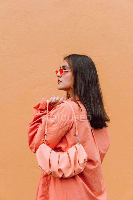 Elegante mujer en traje de moda y bolso a juego y gafas de sol de pie cerca de la pared naranja - foto de stock