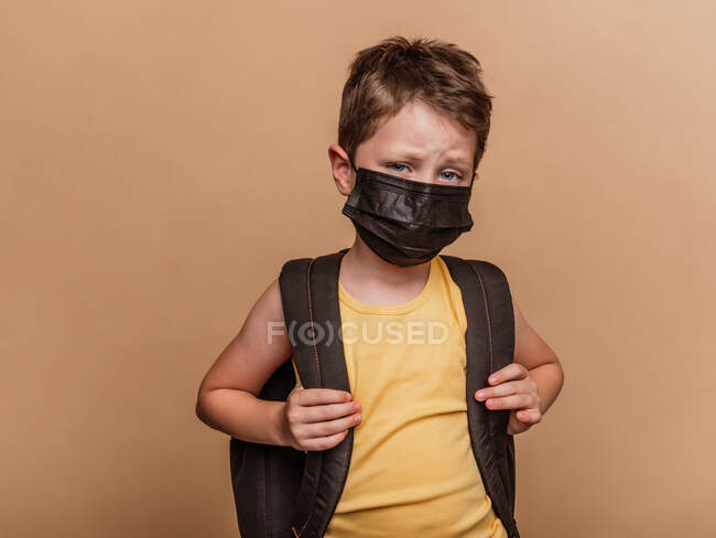Focalizzato preteen scolaro con zaino e in maschera medica protettiva da coronavirus guardando la fotocamera su sfondo marrone in studio — Foto stock
