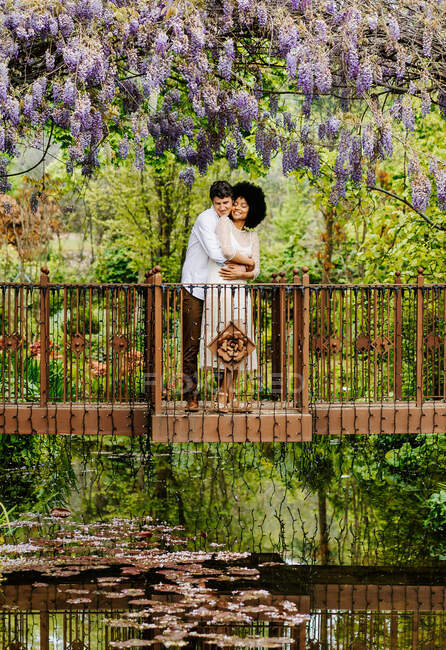 Vue à distance du couple aimant étreignant sur le pont sur l'étang tout en se tenant sous l'arc avec des fleurs de glycine en fleurs dans le jardin naturel — Photo de stock