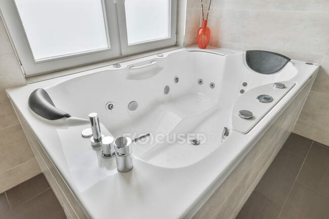 Grand angle de grande baignoire blanche avec des fournitures d'hydromassage situé près de la fenêtre dans la salle de bain luxueuse moderne à la maison — Photo de stock