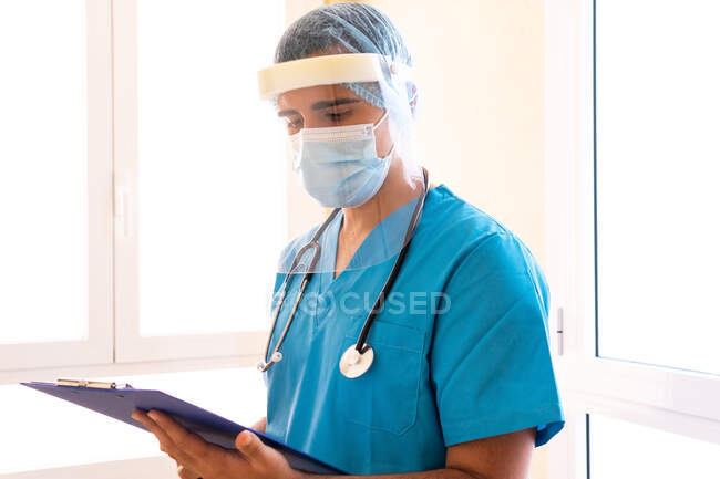 Орієнтований чоловічий медик зі стетоскопом і в масці читання медичного звіту на кишені, стоячи в клініці — стокове фото