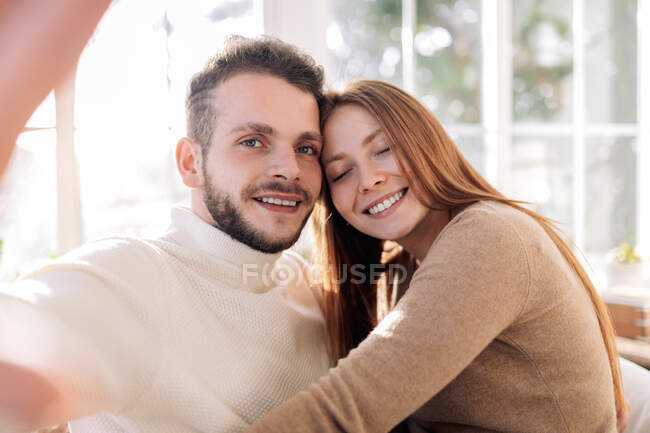 Alegre hombre barbudo con mujer sincera amada tomando autorretrato mientras mira la cámara en casa - foto de stock