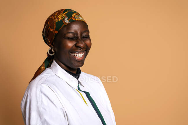 Cheerful African American female model in trendy headscarf and white shirt laughing with closed eyes on beige background in studio — Stock Photo