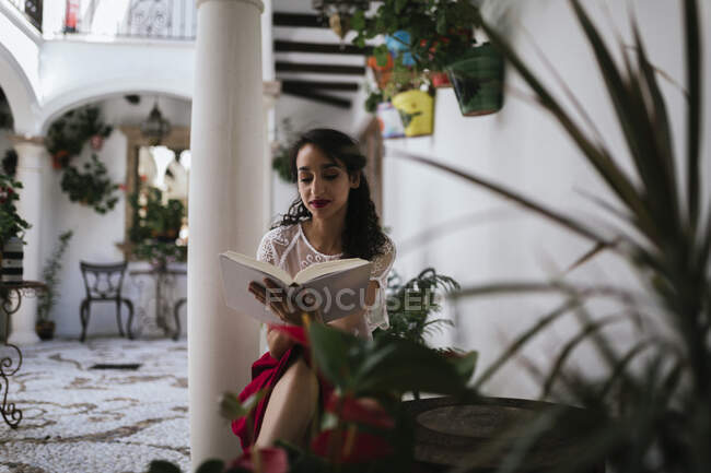 Contenu ethnique féminin en tenue élégante assis sur des tabourets dans le patio et la lecture de roman dans le livre tout en appréciant week-end d'été — Photo de stock