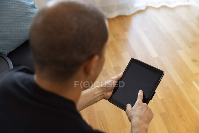 Back view of focused unrecognizable adult male sitting in armchair near table with glass of orange juice and using tablet in morning at home — Stock Photo