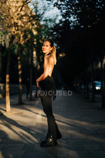 Вид збоку щасливої жінки в модному одязі, що стоїть в місті в сонячний день і дивиться на камеру — стокове фото
