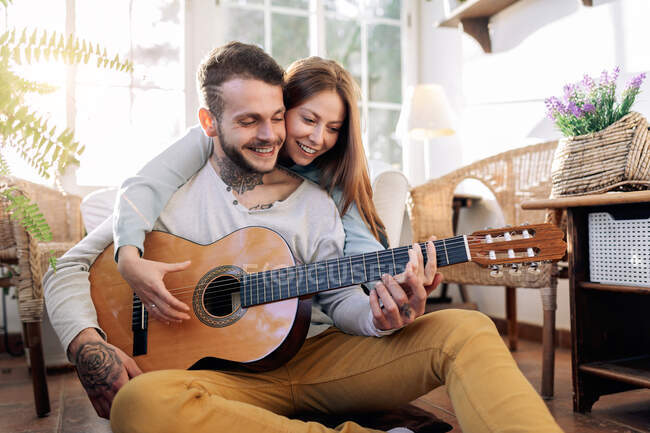 Веселий татуйований чоловічий музикант грає на гітарі біля вмісту коханої жінки, дивлячись один на одного в кріслі в домашній кімнаті — стокове фото