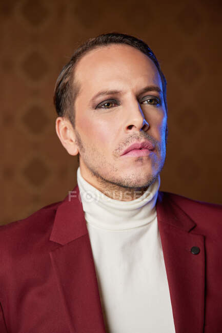 Elegante artista de teatro masculino adulto en chaqueta de color burdeos y con maquillaje mirando hacia otro lado con arrogancia - foto de stock