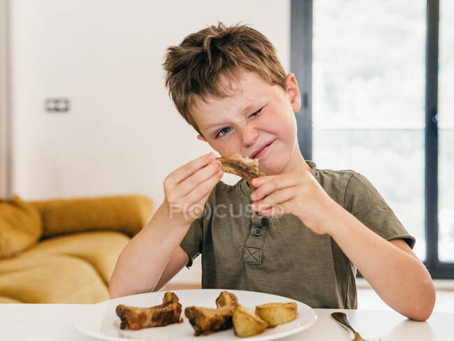Adorable kid eating appetizing pork ribs during lunch at home and winking — Stock Photo