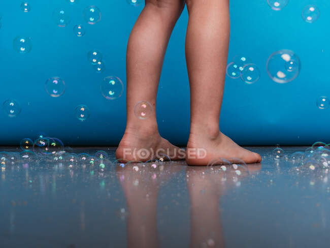 Ernte barfuß Kind steht auf dem Boden im Studio mit fliegenden Seifenblasen auf blauem Hintergrund — Stockfoto
