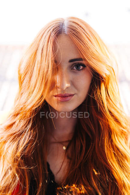 Hübsche Frau mit langen roten Haaren, die vom Sonnenlicht beleuchtet werden und auf verschwommenem Hintergrund in die Kamera schauen — Stockfoto