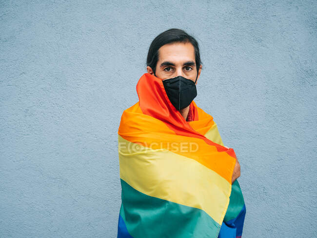 Gay etnico maschio in maschera di protezione e avvolto in arcobaleno LGBT bandiera guardando fotocamera contro grigio muro in città — Foto stock