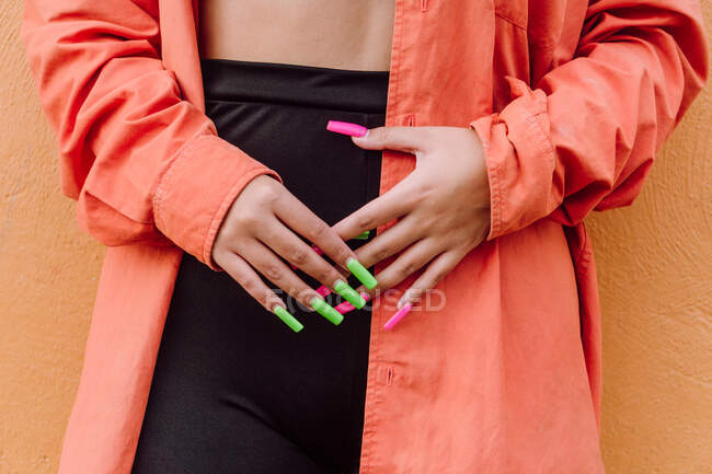Cortar fêmea irreconhecível com unhas compridas coloridas vestindo camisa brilhante em pé perto da parede laranja — Fotografia de Stock