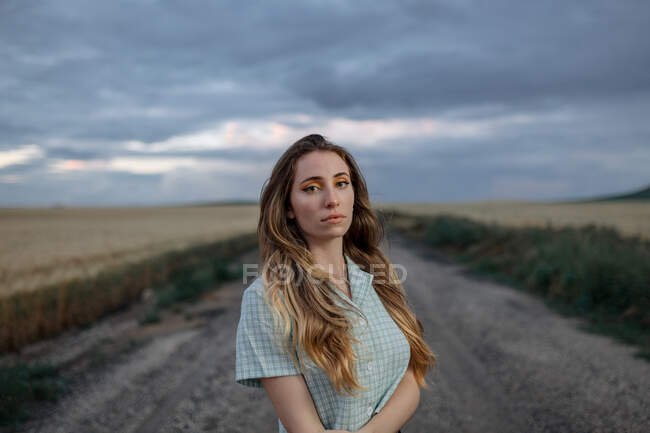 Vue latérale d'une jeune femme consciente regardant une caméra sur une route près d'un pré sous un ciel nuageux en soirée à la campagne — Photo de stock