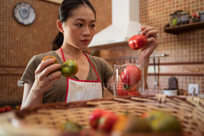 Домохозяйка взвешивает свежие помидоры в стеклянном кувшине на кухонном весе во время приготовления пищи дома — стоковое фото