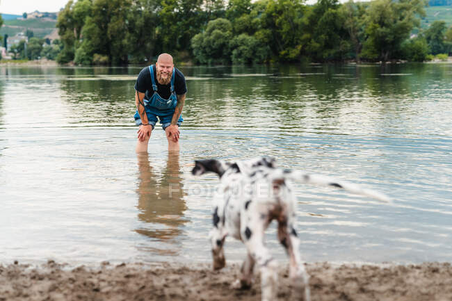 Uomo felice con la barba in abiti casual giocare con il cucciolo mentre in piedi in acqua del lago in estate — Foto stock