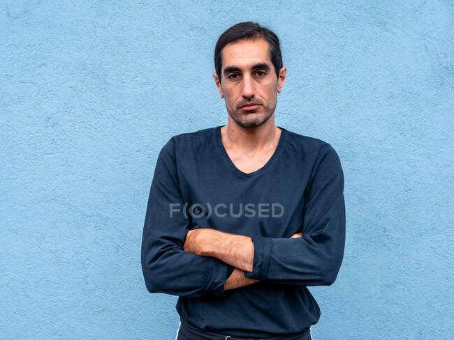 Hombre étnico guapo de pie con los brazos cruzados contra la pared azul claro en la calle de la ciudad y mirando a la cámara - foto de stock
