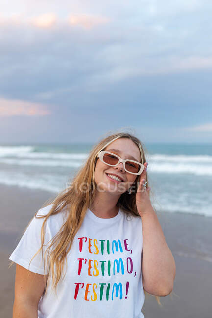 Positive young female in trendy sunglasses and stylish outfit standing at seaside against sea in summer evening — Stock Photo