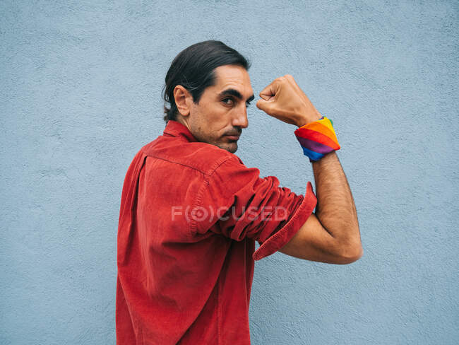 Sereno étnico homossexual masculino com arco-íris bandana na mão mostrando bíceps no fundo da parede cinza na cidade olhando para a câmera — Fotografia de Stock