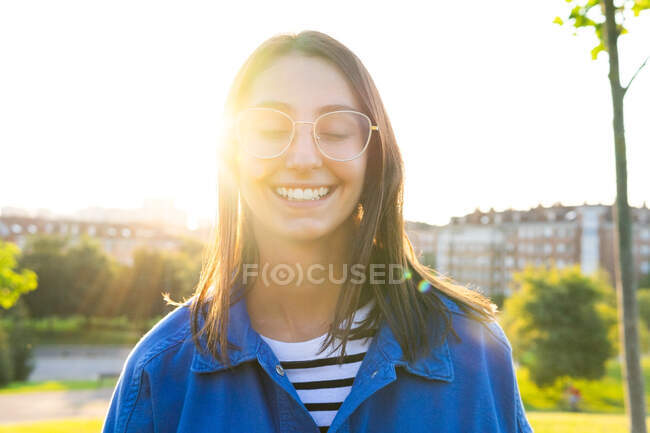 Positive young female in trendy clothes standing with eyes closed smiling on green hill on background of city on sunny day in back lit — Stock Photo