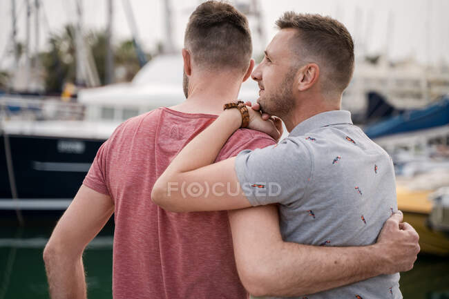 Conteúdo casal de homens homossexuais em t-shirts abraçando enquanto olha para longe no porto — Fotografia de Stock