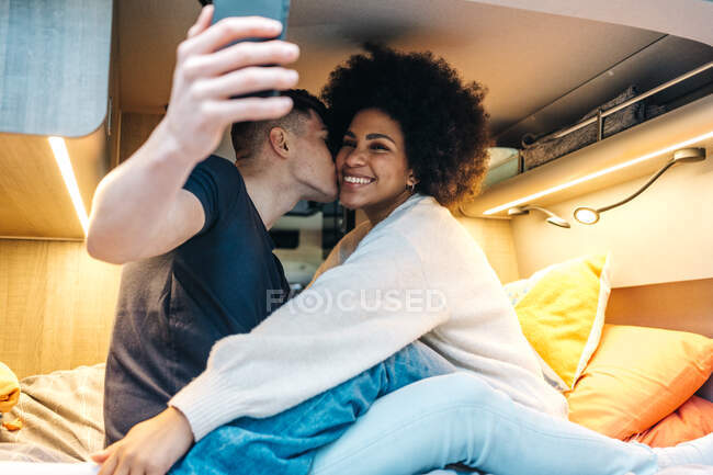 Joven pareja multirracial enamorada tomando selfie en el teléfono inteligente mientras abrazan y besan felizmente dentro de la caravana durante el viaje romántico juntos - foto de stock
