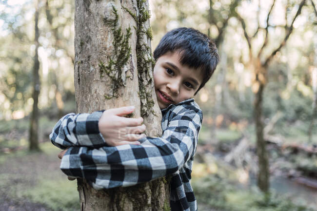 Enfant ethnique sympathique en chemise à carreaux embrassant tronc d'arbre avec mousse et lichen tout en regardant la caméra dans la forêt — Photo de stock